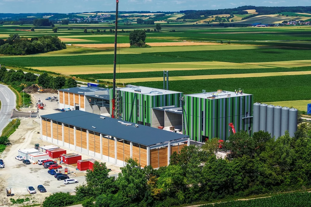 Eine Drohnenaufnahme zeigt die grüne Biomasseanlage in Dingolfing. Ein roter Kran hebt einen Kamin und lässt ihn der Luft schweben. Rechts neben der Biomasseanlage befindet sich eine Lagerhalle, welche teilweise aus Holz gebaut ist.