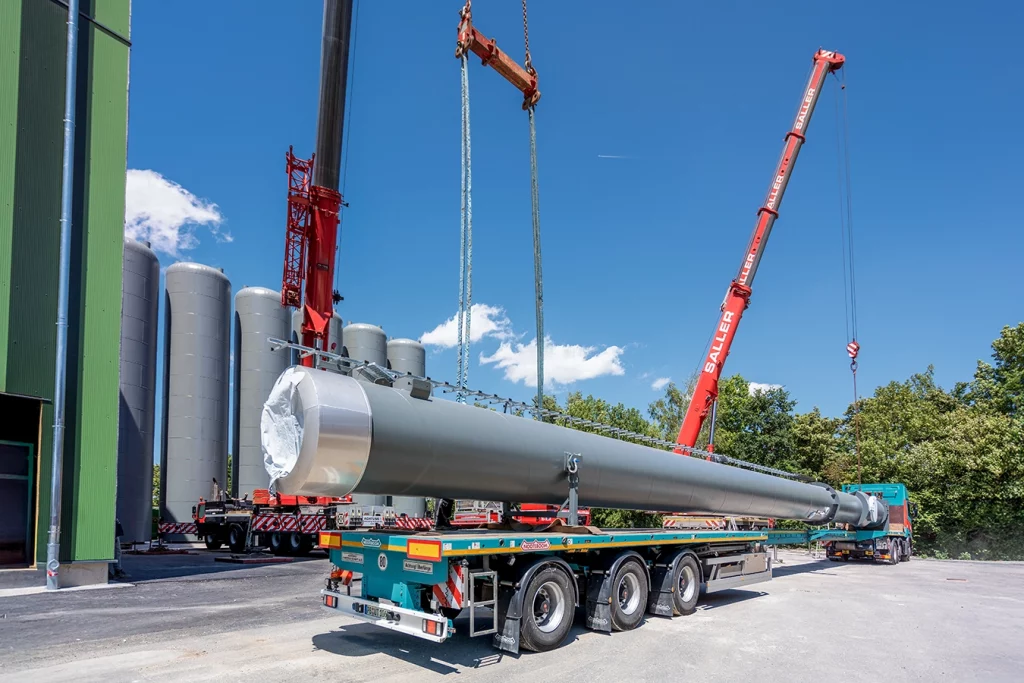 Ein Kamin liegt auf einem blauen LKW. Ein roter LKW dahinter, hat seine Ketten an vorderen Ende des Kamins. Ein weiterer roter LKW nebendran am anderen Ende des Kamins.