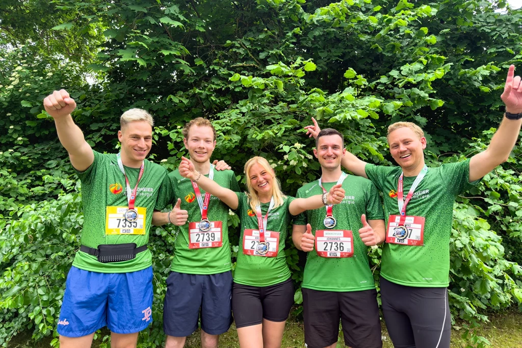 Gruppenbild aus fünf Personen vor einem grünen Gebüsch. Alle fünf Personen tragen ein grünes Sportshirt mit dem Logo von Schmidmeier NaturEnergie. Auf ihrem Shirt befindet sich eine Startnummer, um ihren Hals eine Medaille. Alle fünf Personen jubeln und lachen.
