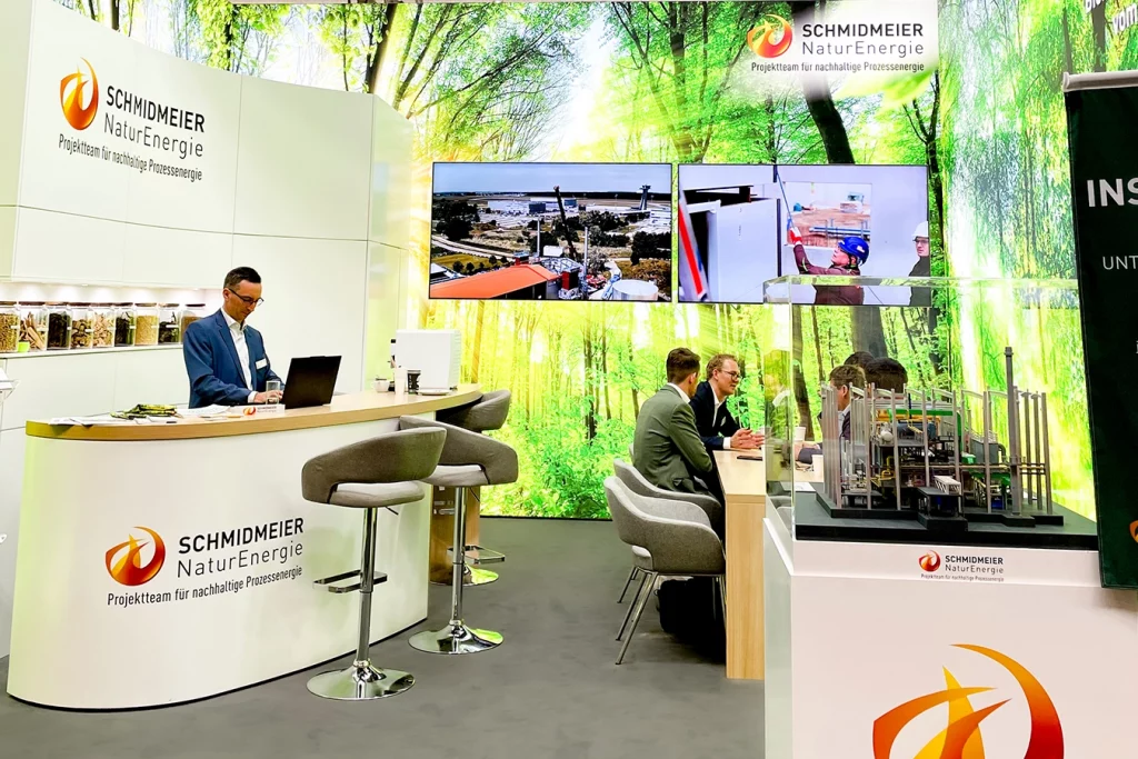 Der Messestand der Schmidmeier NaturEnergie ist im gesamten zu sehen. Links steht ein weißer Stehtisch mit Logo, dahinter ein weißer Schrank. An dem Stehtisch arbeitet ein Mitarbeiter an seinem Laptop. Die Wand zeigt einen grünen Wald mit hellen Strahlen. Rechts am Stand steht ein weiterer Tisch aus Holz, dort sitzen fünf Personen und unterhalten sich. Vor dem Tisch steht ein Modell einer Biomasseanlage.