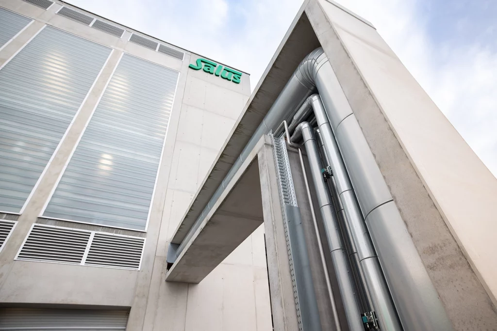 Rohrbrücke einer Biomasseanlage. Mehrere parallel verlaufende metallene Rohre bahn sich waagrecht ihren Weg aus dem Gebäude der Anlage. Nach wenigen Metern verlaufen die Rohre im 90 Grad WInkel nach unten