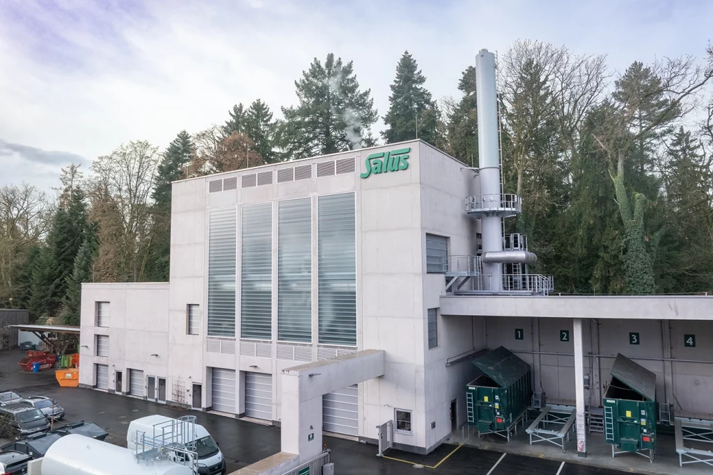 Biomasseanlage in Brückmühl. Die Anlage ist aus Beton errichtet und hat einen Kamin. Im Hintergrund stehen Bäume.