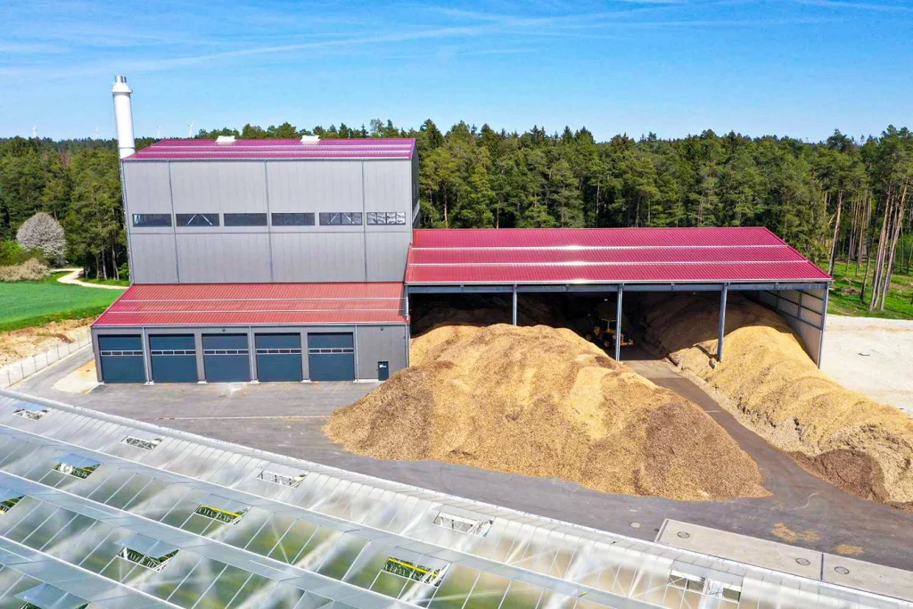 Biomasseanlage in Feulersdorf. Die Anlage hat ein rotes Dach, in der angrenzenden offenen Halle liegt ein riesiger Haufen Hackschnitzel. Vor der Anlage befindet sich ein Gewächshaus.