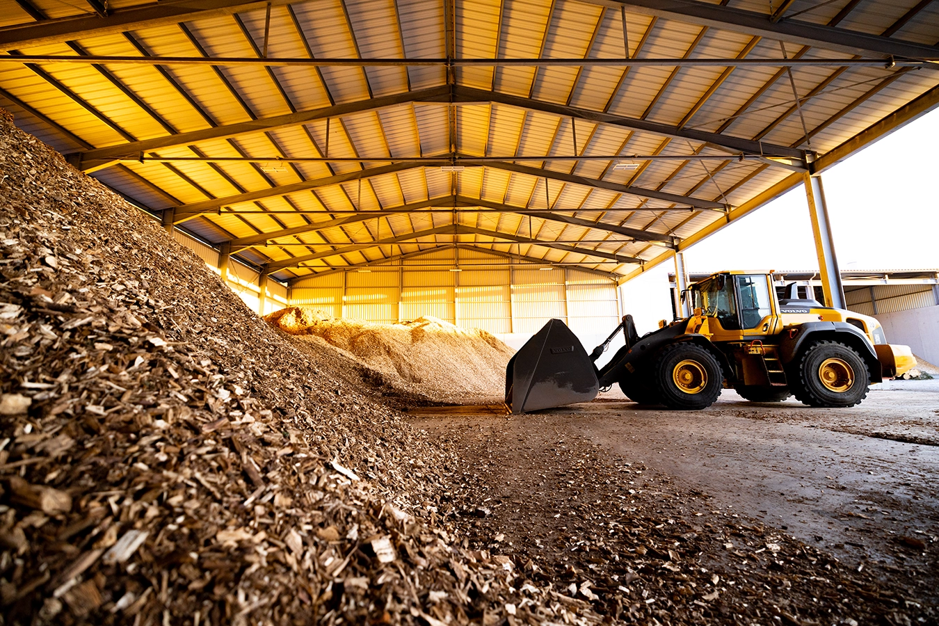 Brennstofflager einer Biomasseanlage in Feulersdorf. In einer großen Halle, zur rechten Seite offen, liegt ein riesiger Haufen Hackschnitzel. Rechts daneben steht ein gelber Bagger.