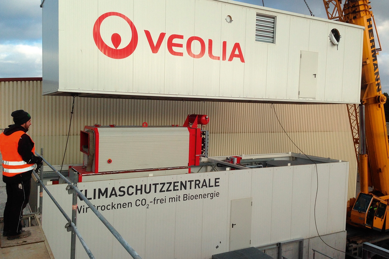 Ein Kran liefert eine Komponente eines Biomasse-Warmwasserkessels. Die Komponente hat die Form eines Containers. Links im Bild steht ein Mitarbeiter mit orangener Sicherheitsweste, der den Vorgang beobachtet. Die Komponente wird auf eine andere Komponente gesetzt.