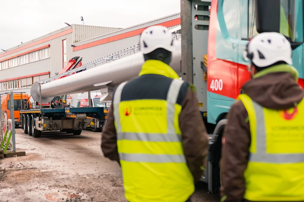 Zwei Mitarbeiter von Schmidmeier mit dem Rücken zur Kamera stehend. Ausgerüstet mit Sicherheitskleidung. Gelbe Warnweste mit Schmidmeier-Aufschrift und Sicherheitshelm. Beobachten die Anlieferung der Montagekomponenten in Hüllhorst.