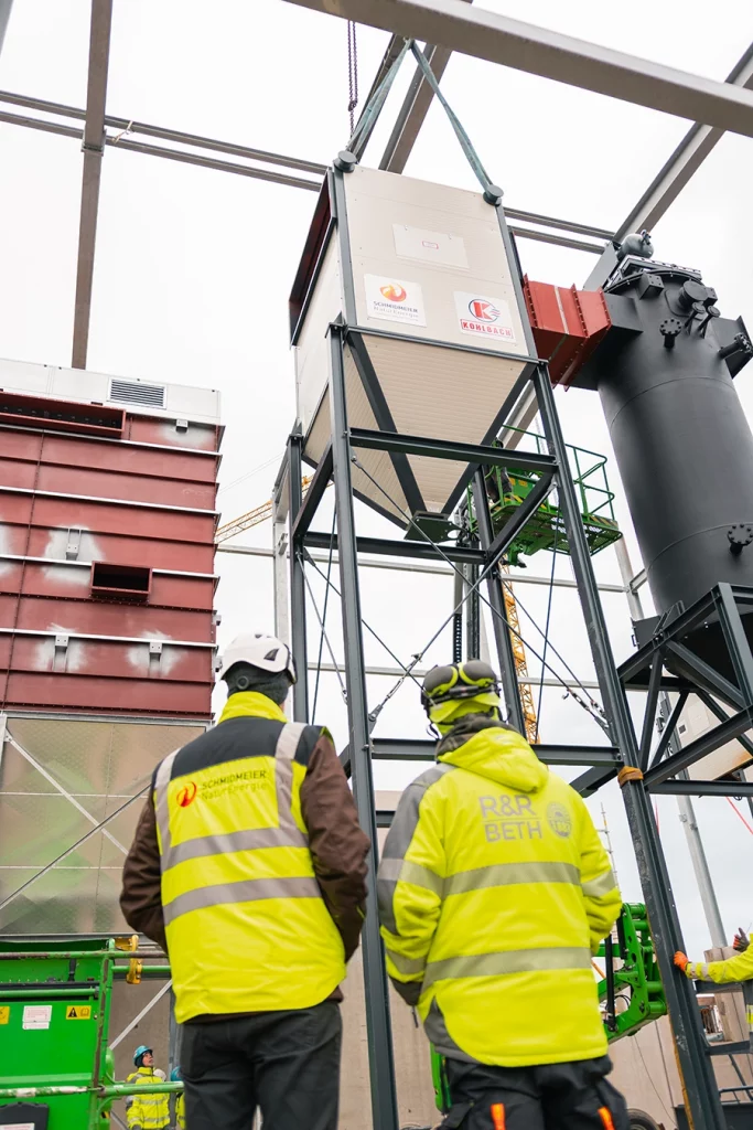 Zwei Bauarbeiter stehen mit gelber Sicherheitsweste und Helm vor einer Komponente einer Biomasseanlage, die gerade von einem Kran heruntergelassen wird.