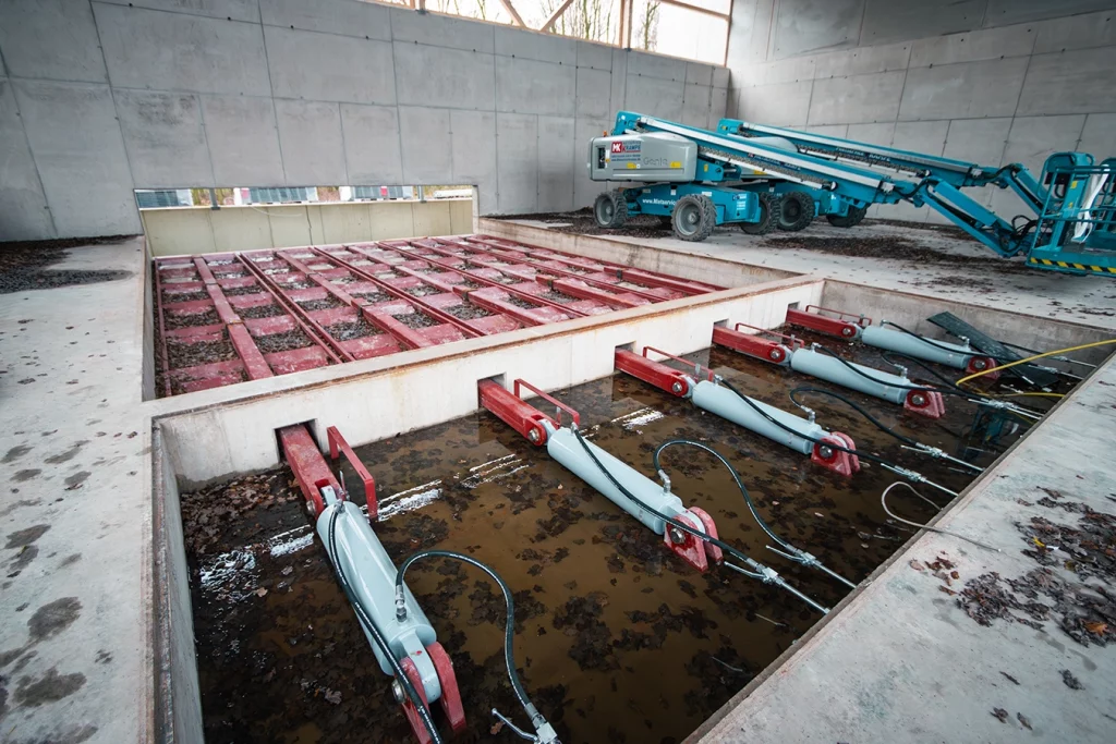 Das Bild zeigt das Innere einer offenen Industriehalle aus Beton. In einem rechteckigen, eingelassenen Becken befinden sich mehrere rote Metallrahmen mit angeschlossenen grauen Hydraulikzylindern, die teilweise im Wasser liegen. Im Hintergrund steht eine türkisfarbene Hebebühne auf der Betonfläche. Hohe Wände und schmale Fensteröffnungen lassen Tageslicht in die Halle.
