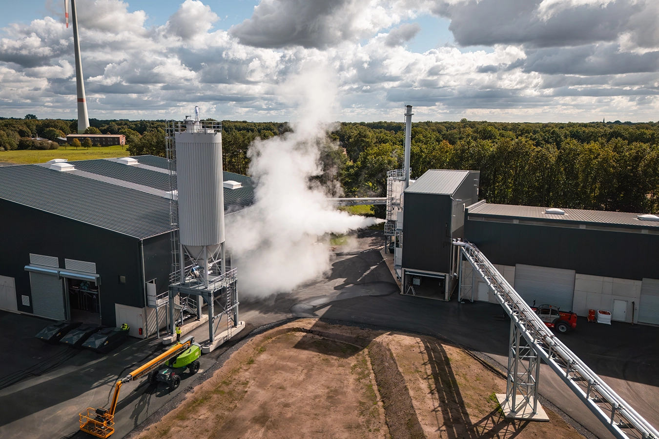 Auf einem geteerten Industriegelände steht ein Biomassekessel in Verbindung mit schwarzen Gebäuden. Aus einem Dampfrohr schießt Dampf in die Luft.