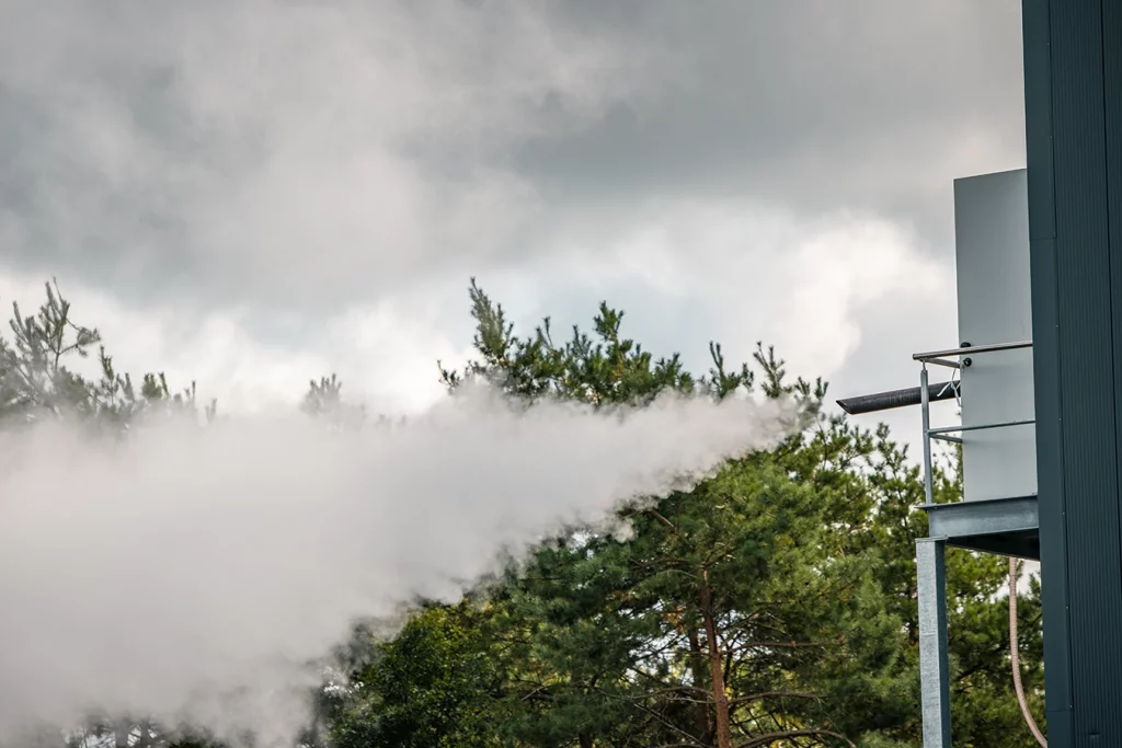 Dampf schießt aus einer Dampfleitung eines Biomassekessels. Im Hintergrund sieht man grüne Bäume.