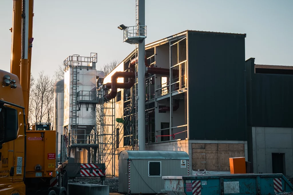 Baustelle eines Biomassekessels. Auf der linken Seite befinden sich Komponenten im freien die durch Rohre in ein nebenstehendes, noch offenes Gebäude, führen.