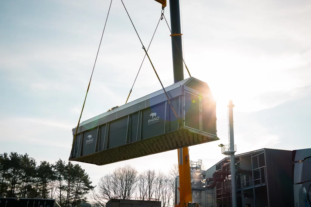 Ein Kran transportiert einen Komponente eines Biomassekessels. Die Komponente hat eine schwarze Außenfassade.