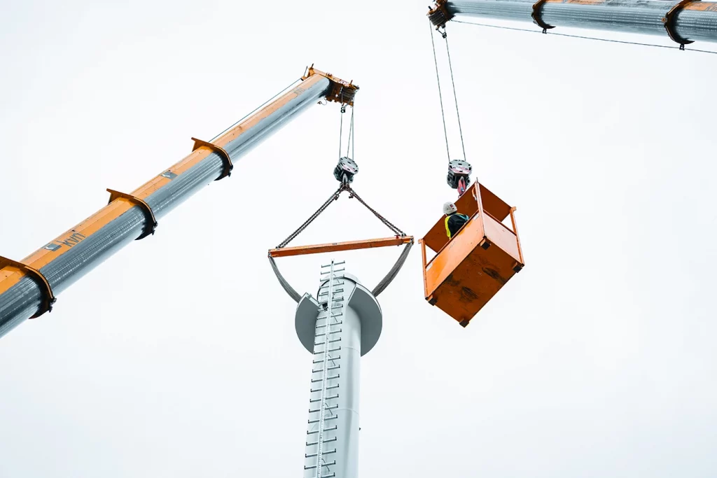 Ein Kran hebt einen weißen Kamin. Daneben befindet sich ein weiterer Kran, welcher eine orangene Kabine mit einem Mitarbeiter trägt. Der Mitarbeiter beobachtet den Vorgang.