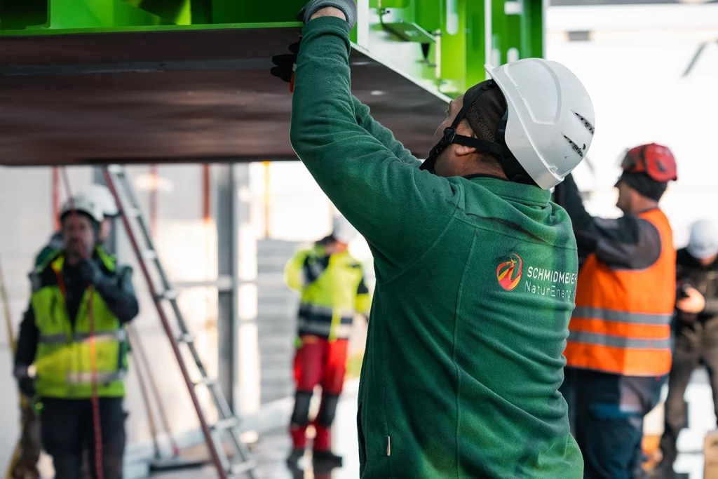 Ein Mitarbeiter der Schmidmeier NaturEnergie mit grüner Stoffjacke und weißem Sicherheitshelm hilft beim Transport einer Komponente. Diese wird von einem Kran in der Luft getragen. Im Hintergrund sind weitere Mitarbeiter.