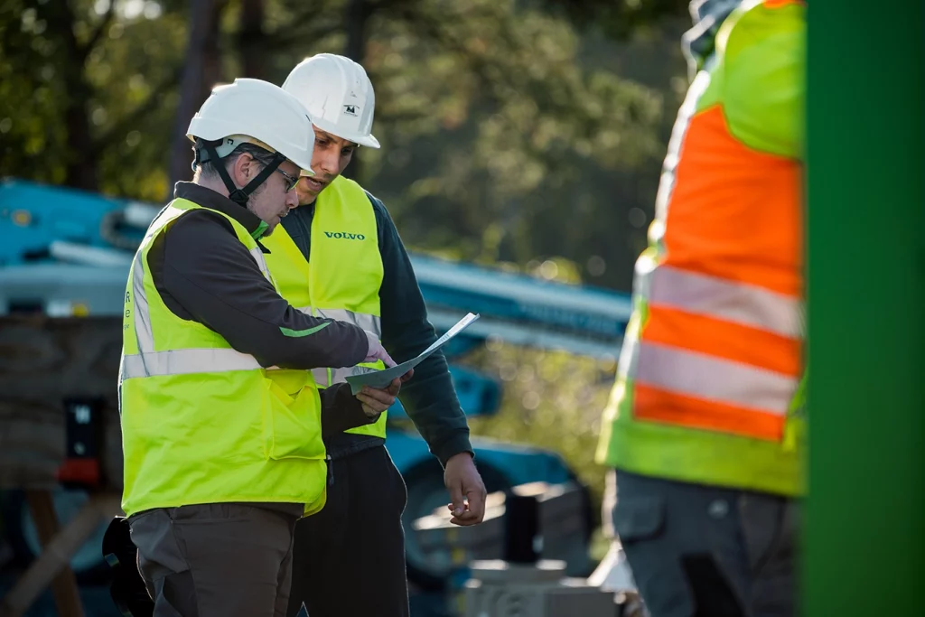 Zwei Mitarbeiter der Schmidmeier NaturEnergie mit Sicherheitsweste unterhalten sich miteinander. Einer der beiden zeigt dem anderen etwas auf einem Blatt Papier. Außerdem tragen sie weiße Sicherheitshelme.