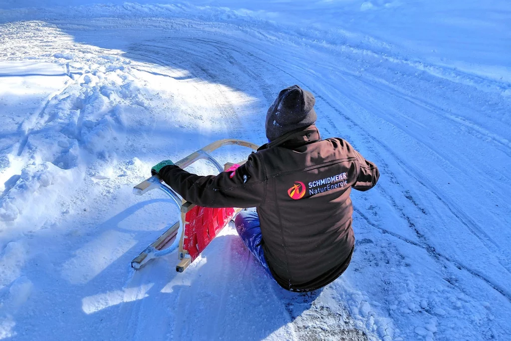Man sieht einen Mitarbeiter der Schmidmeier NaturEnergie von hinten am Boden im Schnee sitzen. Er trägt eine Jacke mit dem Logo der Schmidmeier NaturEnergie und hält einen Schlitten in den Händen.