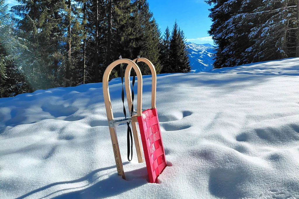 Ein roter Schlitten aus Holz steckt im Schnee. Im Hintergrund sieht man Bäume und es scheint die Sonne.