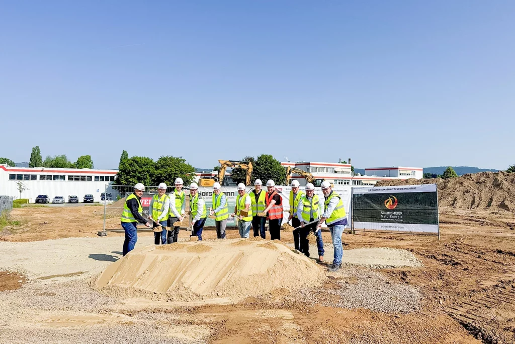 Eine Gruppe von Männern steht mit Schaufel und Warnweste vor einem Haufen Erde. Sie tragen alle Sicherheitshelme. Im Hintergrund stehen zwei gelbe Bagger.
