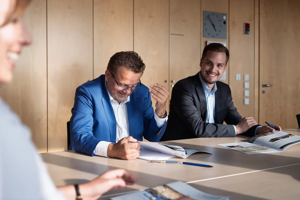 Thomas Schmidmeier und David Dluhosch in einem Besprechungsraum an einem Tisch sitzend, vor ihnen Prospekte