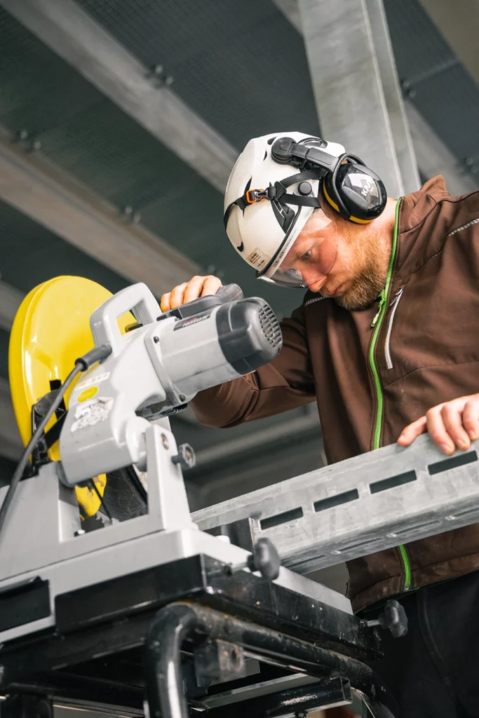 Ein Bauerbeiter sägt ein Metallstück mit einer Kreissäge. Der Bauerbeiter hat einen blonden Bart und weißen Helm mit Kopfhörern auf.