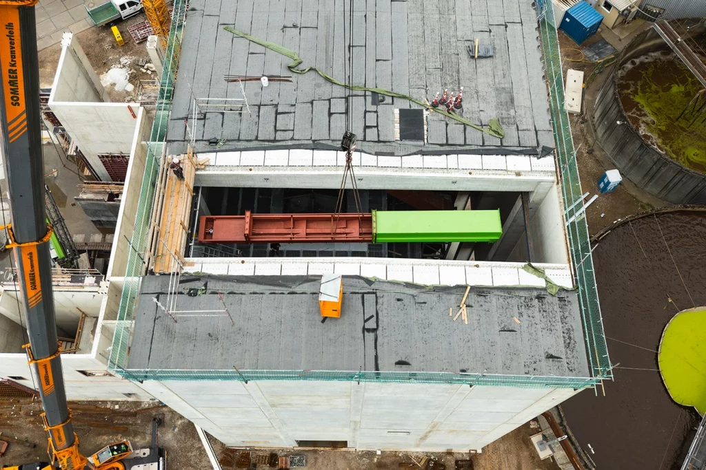 Ein Kran lässt einen Querförderer hinab in einen Biomasseanlage. Der Vorgang ist aus der Vogelperspektive zu beobachten.