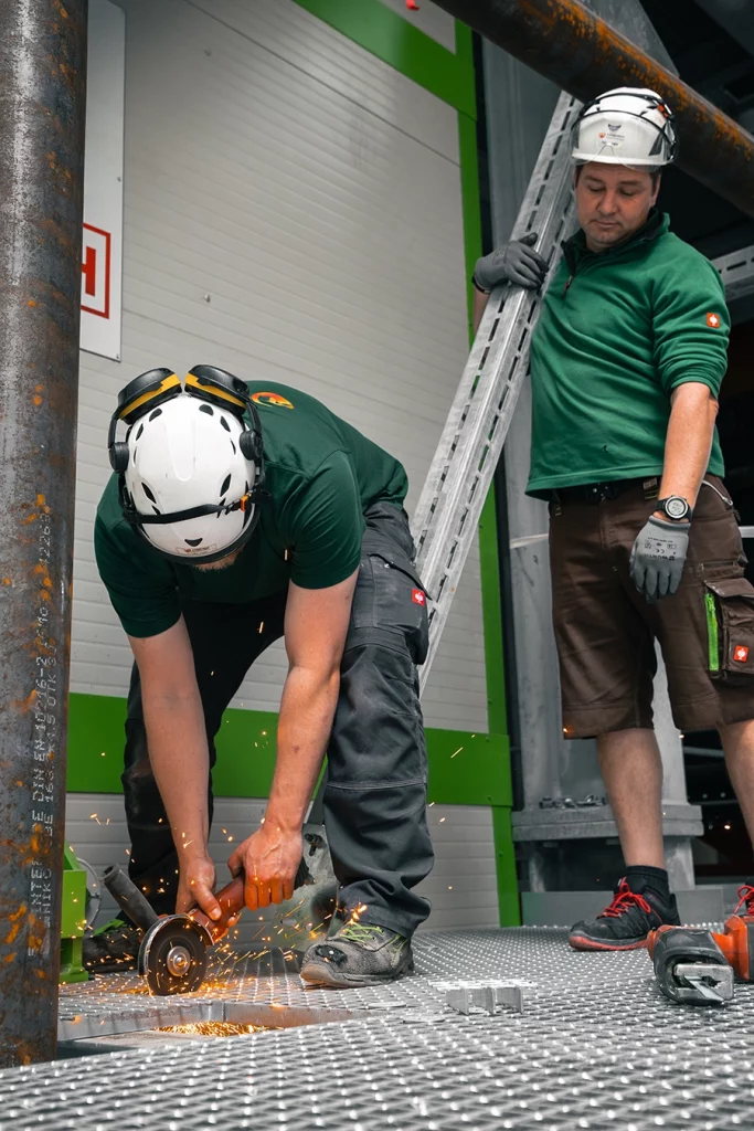 Ein Bauerbeiter steht gebückt und sägt ein Stück Metallgitter aus dem Boden. Ein weiterer Bauarbeiter steht direkt hinter, dieser hält ein längliches Metallstück in der Hand, welches er auf dem Boden abstützt.