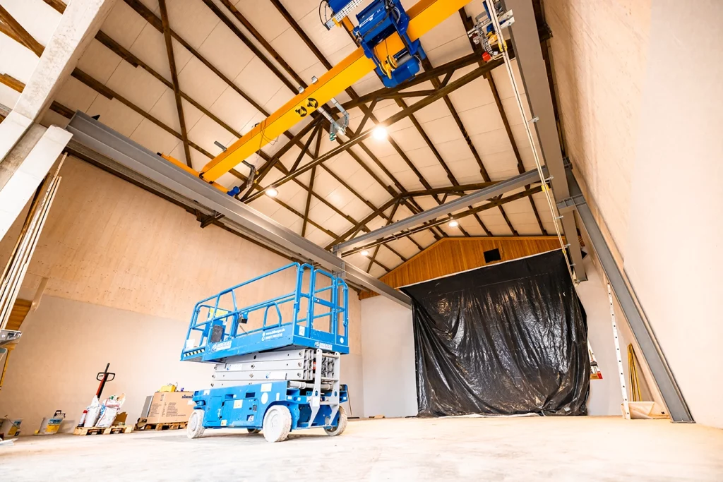 Eine große leere Halle mit flachem Spitzdach. Am Eingang befindet sich eine große schwarze Plane, die diesen verdeckt. In der Mitte der Halle befindet sich eine blaue Hubarbeitsbühne. Die Halle wird zu einer Werkstatt für die Schmidmeier NaturEnergie umgebaut.