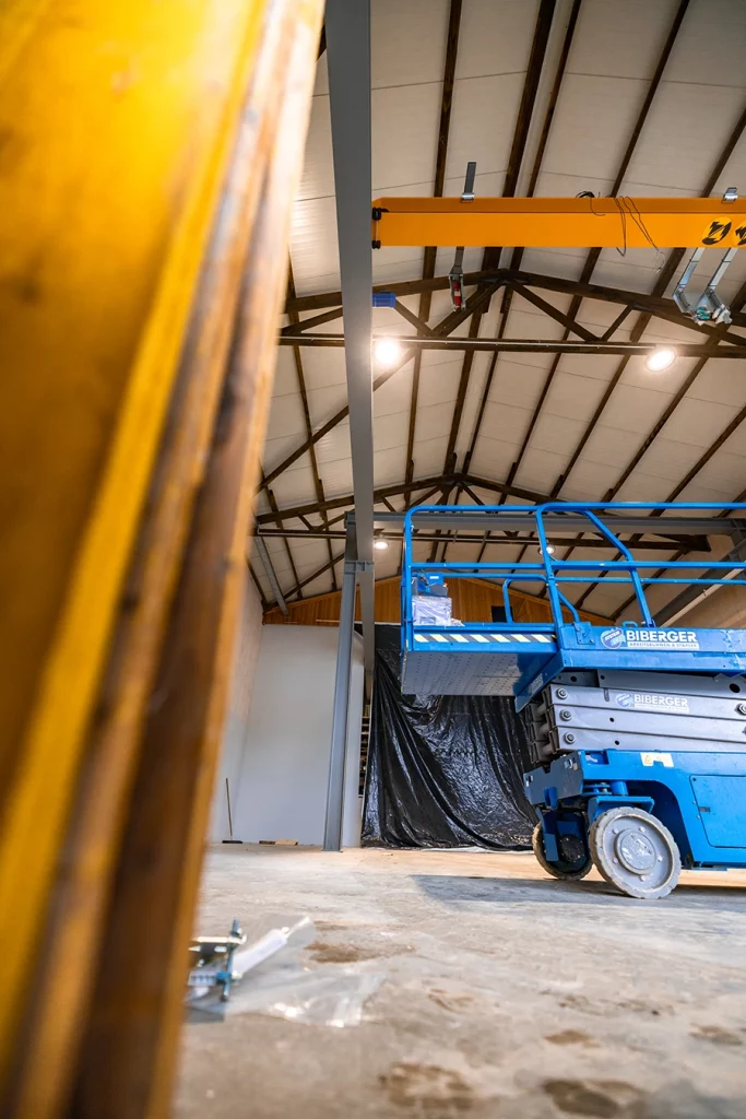 Eine große leere Halle mit flachem Spitzdach. Am Eingang befindet sich eine große schwarze Plane, die diesen verdeckt. In der Mitte der Halle befindet sich eine blaue Hubarbeitsbühne. Die Halle wird zu einer Werkstatt für die Schmidmeier NaturEnergie umgebaut.