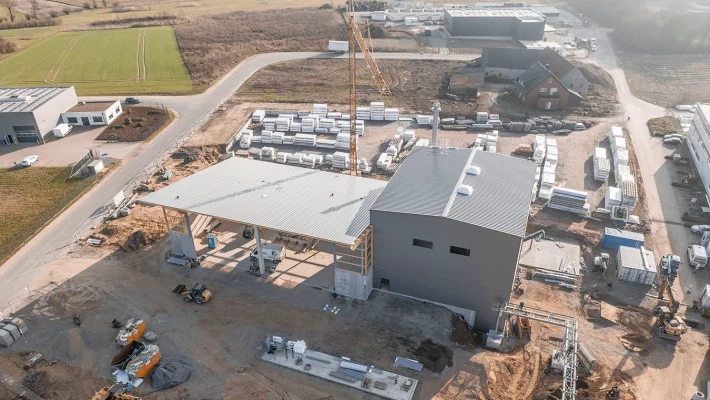 Drohnenaufnahme einer Baustelle der Biomasseanlage in Hüllhorst. Der Boden ist mit Erde bedeckt, auf dem mehrere Bagger sowie ein Kran steht. In der Mitte befindet sich die Anlage, ein dunkelgraues viereckiges Gebäude mit einer Halle die links davon anschließt.