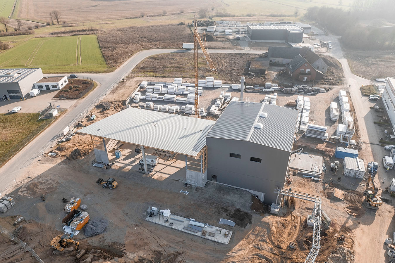 Drohnenaufnahme einer Baustelle der Biomasseanlage in Hüllhorst. Der Boden ist mit Erde bedeckt, auf dem mehrere Bagger sowie ein Kran steht. In der Mitte befindet sich die Anlage, ein dunkelgraues viereckiges Gebäude mit einer Halle die links davon anschließt.
