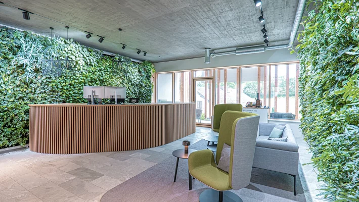 Empfangsbereich des Büros der Schmidmeier NaturEnergie in Zeitlarn. Die Wände sind mit grünen Pflanzen bedeckt. Auf der linken Seite befindet sich ein großer Tisch mit einer Holzfassade. Auf der rechten Seite steht eine graue Couch und zwei grau/grüne Stühle.