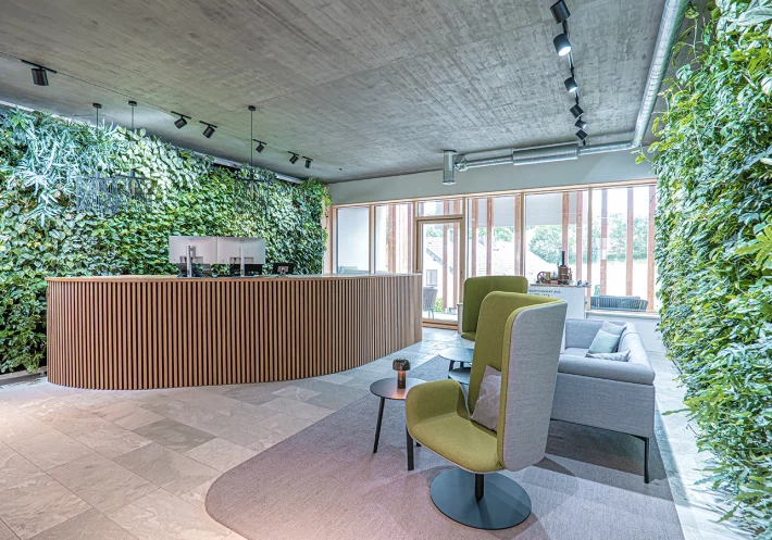 Empfangsbereich des Büros der Schmidmeier NaturEnergie in Zeitlarn. Die Wände sind mit grünen Pflanzen bedeckt. Auf der linken Seite befindet sich ein großer Tisch mit einer Holzfassade. Auf der rechten Seite steht eine graue Couch und zwei grau/grüne Stühle.