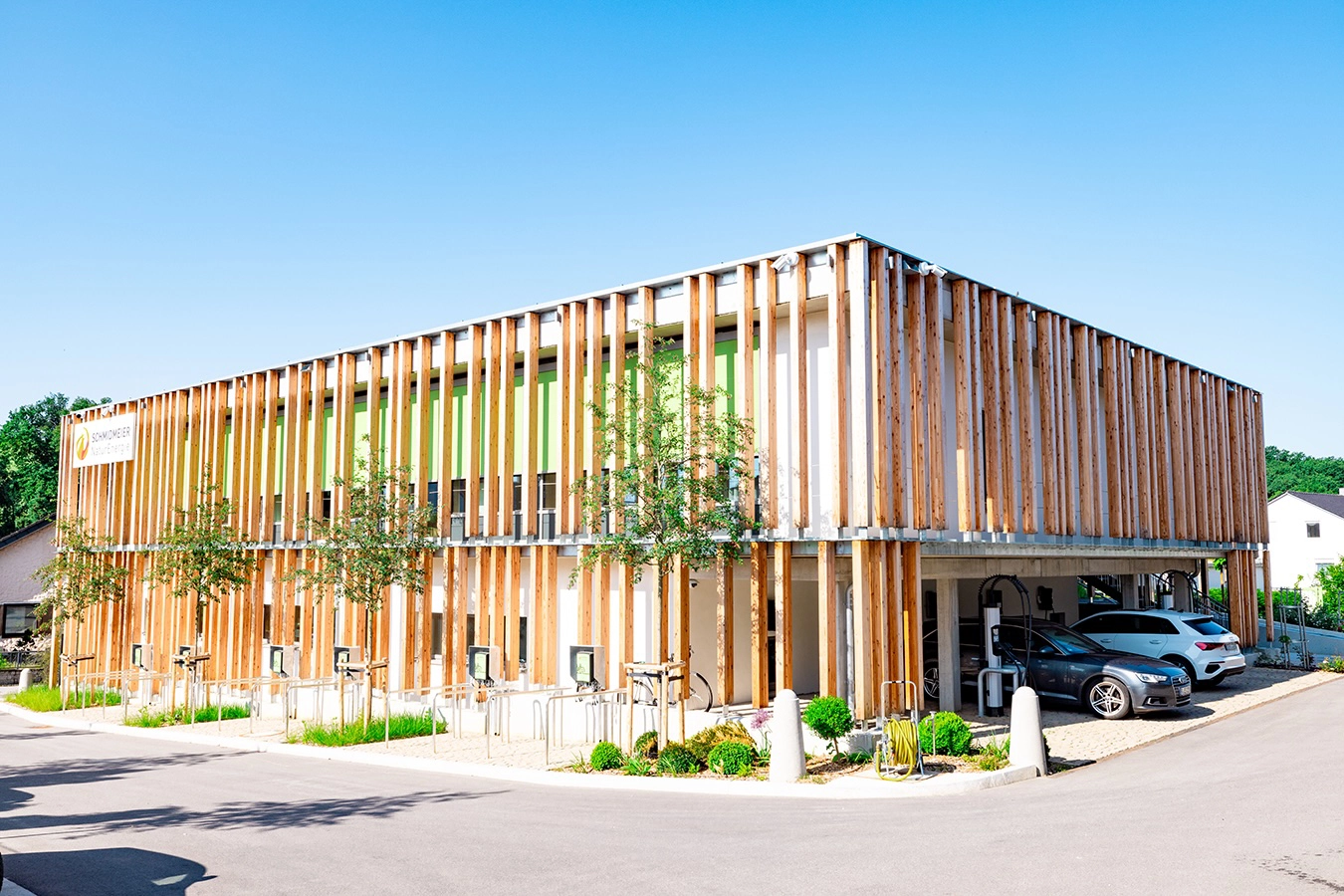 Verwaltungsgebäude von der Ostseite, helle Holzfassade, junge Bäume im Vordergrund, zwei Autos stehen am Parkplatz