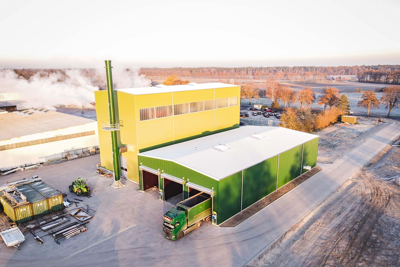 Ansicht der Anlage mit Bennstofflager von außen / oben und rauchendem Kamin. Im Hintergrund Landschaft mit Schnee bedeckt.