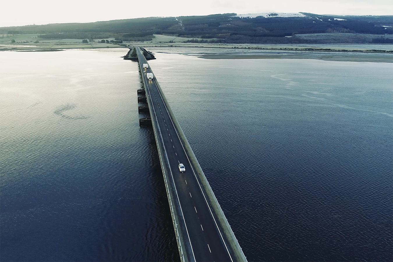 Ansicht des Schwertransportes der Anlieferung der Baukomponenten. Drohnenaufnahme. Fahrzeuge befinden sich auf Brücke über Wasser/Meer.