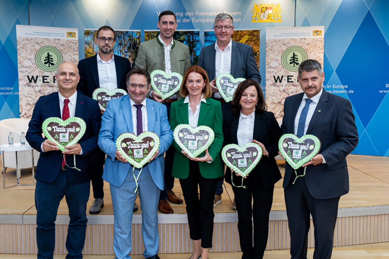 Dieses Foto zeigt eine Gruppe von Menschen, die auf einer Bühne mit herzförmigen Schildern posieren, während im Hintergrund Werbeplakate für Bayern in Europe zu sehen sind. Die Veranstaltung fand im Rahmen einer Initiative zur Stärkung der Holzenergie in Europa statt.